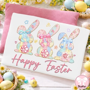 Könnte beinhalten: Weißes, österliches Dekokissen mit drei Patchwork-Hasen mit Blumenmustern, Schleifen und Blumenakzenten. Die Worte "Happy Easter" sind in rosa Schrift unten geschrieben.