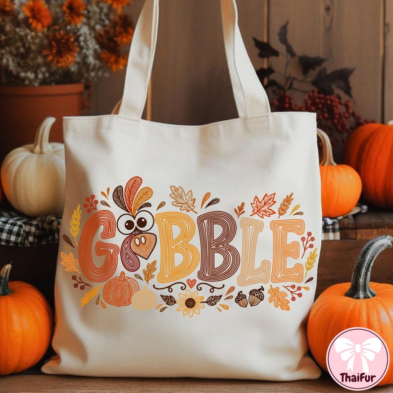May include: A cream-colored canvas tote bag featuring a graphic print with the word GOBBLE in large, multi-colored, striped letters. The letter O is replaced by a turkey face, surrounded by illustrations of autumn leaves, pumpkins, and acorns.