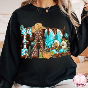 May include: Black sweatshirt with a colorful "FALL" graphic. The design features a cowboy hat, turquoise cowboy boots, a pumpkin, and floral accents. The letter "F" has a blue and white checkered pattern, and the letter "L" has a brown pattern.