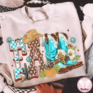 May include: Cream-colored sweatshirt with a graphic design featuring the word "FA" in a Western style. The design includes a cowboy hat, turquoise cowboy boots, a pumpkin, and a cactus. The letters are decorated with a checkered pattern and a brown pattern.