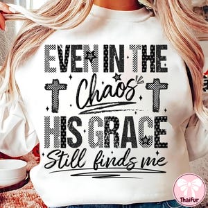 May include: White sweatshirt with the text "EVEN IN THE Chaos HIS GRACE Still finds me" in black. The text includes cross and star graphics. The sweatshirt has a crew neck.