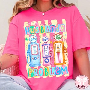 Pode incluir: Uma t-shirt rosa brilhante com a frase "That's my Husband's Problem" em letras coloridas e divertidas. O design inclui ilustrações de bombas de gasolina vintage em azul, rosa e amarelo, com a palavra "GAS" em cima.