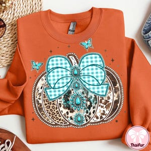May include: Orange sweatshirt featuring a pumpkin design with a turquoise and white gingham bow, turquoise gemstone accents, and a leopard print center. Three turquoise butterflies and star accents complete the design.