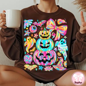 May include: Brown sweatshirt featuring a colorful Halloween design. The design includes stacked jack-o'-lanterns, ghosts, bats, stars, and a large bow. The artwork uses bright neon colors, creating a festive and eye-catching look.