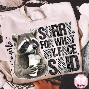 May include: A light pink sweatshirt featuring a raccoon holding a drink with a straw. The text "SORRY FOR WHAT MY FACE SAID" is printed in a mix of checkered, leopard print, and solid black fonts. Stars and graphic elements surround the raccoon.