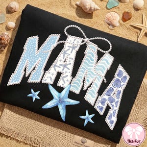 May include: Black t-shirt with the word "MAMA" in a decorative font. The letters are adorned with blue patterns, including polka dots, starfish, and seashells. A string of pearls connects the two "A" letters, and several blue starfish are scattered around the word.