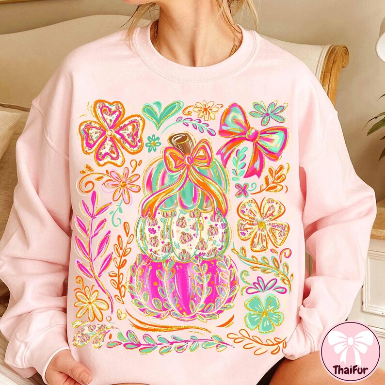 May include: A light pink sweatshirt featuring a stacked pumpkin design with a bow and floral accents in shades of pink, orange, green, and blue. The design includes a variety of flowers and leaves. The sweatshirt is a comfortable, casual style.