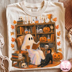 May include: A cream-colored t-shirt featuring a graphic of a ghost sitting in an armchair reading a book and holding a mug. The scene includes a bookshelf filled with pumpkins, skulls, candles, and books, with a black cat and autumn leaves.