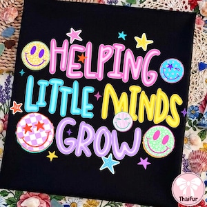 May include: A black t-shirt featuring the text HELPING LITTLE MINDS GROW in pink, blue, yellow, and purple. The design includes various colorful smiley faces with patterns like leopard print, checkers, and stripes, along with small star icons.