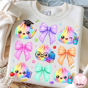 May include: A cream-coloured sweatshirt featuring a colourful graphic print of five rainbow-coloured, glittery, smiling dumpling characters. The design includes four large glittery bows in purple, pink, orange, and teal, along with school supplies.