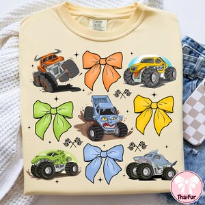 K&ouml;nnte beinhalten: Hellgelbes T-Shirt mit Cartoon-Monstertrucks in verschiedenen Farben und Schleifen. Das T-Shirt hat einen Rundhalsausschnitt und kurze &Auml;rmel. Der Text "Comfort Colors" ist auf dem Kragen zu sehen. Die Monstertrucks sind orange, gelb, gr&uuml;n und blau.