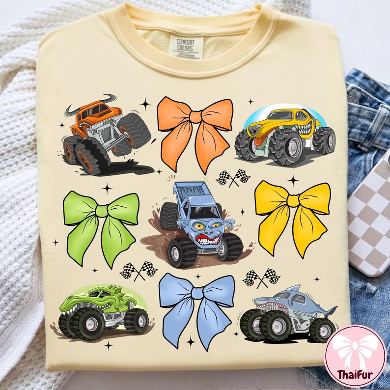 K&ouml;nnte beinhalten: Hellgelbes T-Shirt mit Cartoon-Monstertrucks in verschiedenen Farben und Schleifen. Das T-Shirt hat einen Rundhalsausschnitt und kurze &Auml;rmel. Der Text "Comfort Colors" ist auf dem Kragen zu sehen. Die Monstertrucks sind orange, gelb, gr&uuml;n und blau.