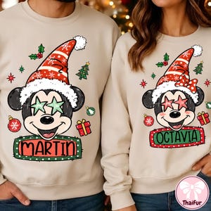 Könnte beinhalten: Zwei beige Sweatshirts mit Rundhalsausschnitt und einer Cartoon-Maus mit Weihnachtsmütze und sternförmiger Brille. Das linke Sweatshirt zeigt den Namen MARTIN in einem grünen Kasten, das rechte Sweatshirt den Namen OCTAVIA.