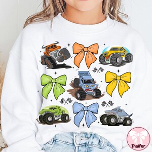 K&ouml;nnte beinhalten: Wei&szlig;es Sweatshirt mit bunten Monstertruck-Grafiken und dekorativen Schleifen. Die Monstertrucks sind in Orange, Gelb, Gr&uuml;n und Blau gehalten und haben gro&szlig;e Reifen und aggressive Designs. Die Schleifen sind orange, gr&uuml;n, gelb und blau.