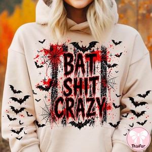 May include: A beige hooded sweatshirt featuring a graphic print with the text BAT SHIT CRAZY in black, distressed lettering. The design includes red paint splatters, black spiderwebs, and numerous small black bat silhouettes across the front.