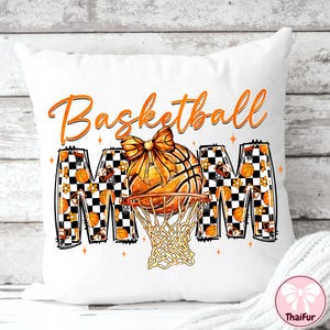 May include: White decorative pillow with the words "Basketball Mom" in orange script. The design features a basketball with a bow, a net, and the letters "MOM" with basketball and floral patterns.