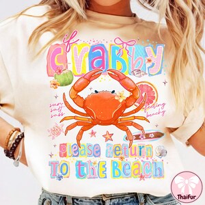May include: A cream-coloured t-shirt featuring a vibrant crab graphic and the phrase "If Crabby, Please Return To The Beach." The crab is orange with yellow flowers. The design incorporates beach-themed illustrations.