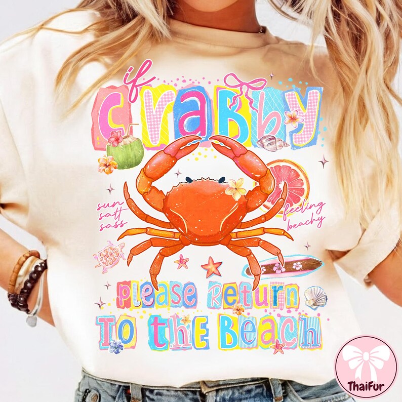 May include: A cream-coloured t-shirt featuring a vibrant crab graphic and the phrase "If Crabby, Please Return To The Beach." The crab is orange with yellow flowers. The design incorporates beach-themed illustrations.