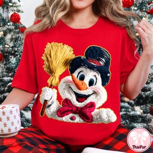 May include: A red t-shirt with a snowman graphic. The snowman has a black hat, orange nose, and a red bow tie. He is holding a broom. The shirt is part of a Christmas theme.