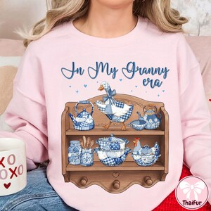 K&ouml;nnte beinhalten: Hellrosa Sweatshirt mit dem Schriftzug "In My Granny era" in blauer Schrift. Ein Holzregal zeigt blau-wei&szlig; kariertes K&uuml;chengeschirr, darunter eine Teekanne, eine Gans und ein Huhn.