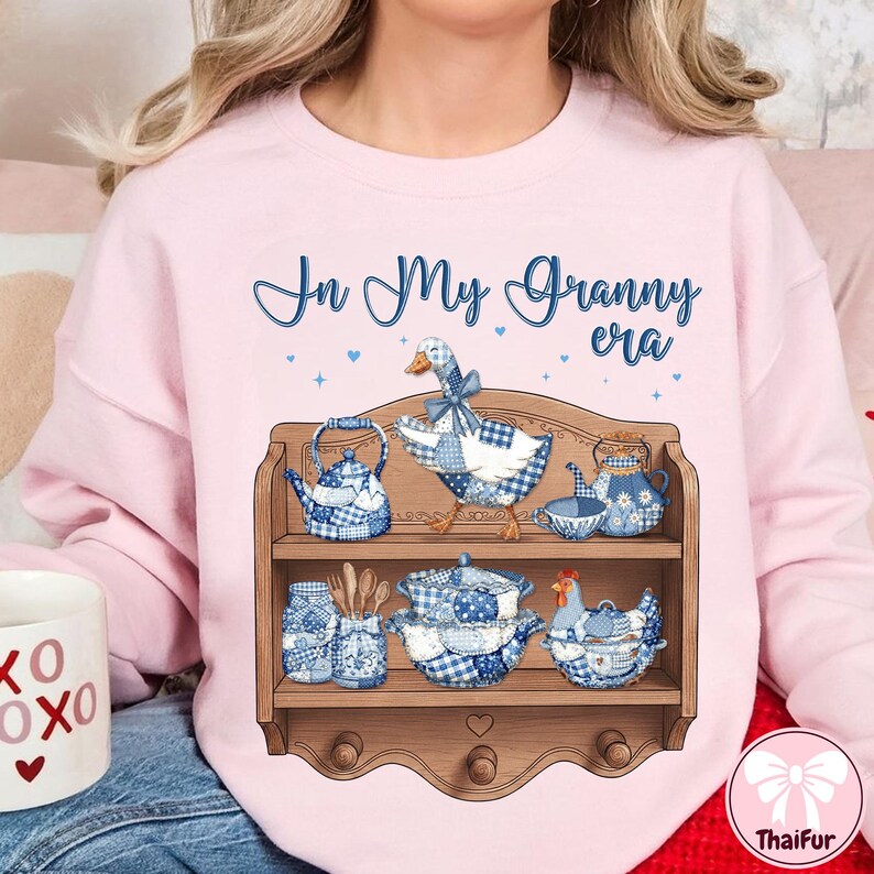 K&ouml;nnte beinhalten: Hellrosa Sweatshirt mit dem Schriftzug "In My Granny era" in blauer Schrift. Ein Holzregal zeigt blau-wei&szlig; kariertes K&uuml;chengeschirr, darunter eine Teekanne, eine Gans und ein Huhn.