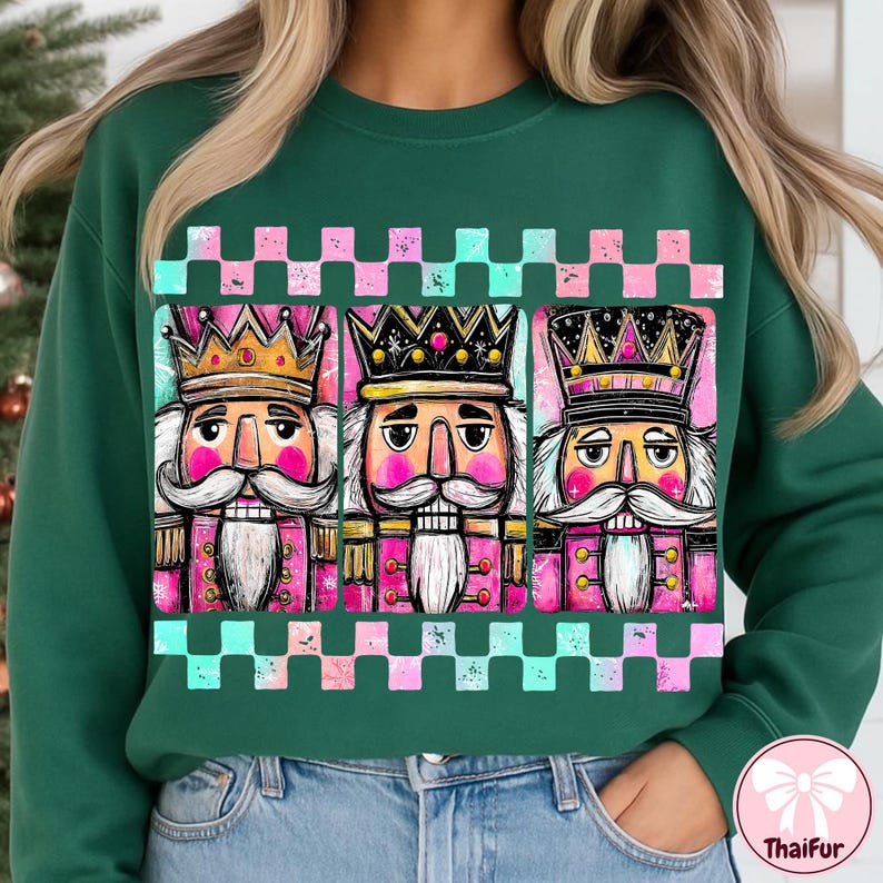K&ouml;nnte beinhalten: Dunkelgr&uuml;nes Sweatshirt mit einer Grafik von drei Nussknacker-Figuren. Jeder Nussknacker hat eine Krone, einen wei&szlig;en Schnurrbart und eine rosa-goldene Uniform. Die Grafik wird von einem karierten Muster in Rosa und T&uuml;rkis umrahmt.