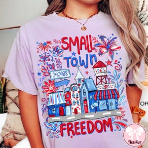 May include: A lilac-colored t-shirt featuring a colorful graphic with the words "SMALL TOWN FREEDOM." The design includes illustrations of buildings, flowers, and a water tower in red, white, and blue.