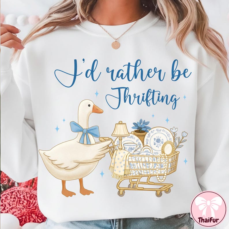 May include: White sweatshirt with the text "I'd rather be Thrifting" in blue script. A cartoon goose with a blue bow tie pushes a shopping cart filled with vintage dishes, a lamp, and a pearl necklace.