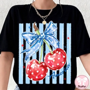 May include: Black t-shirt featuring a patriotic design with two red cherries adorned with white stars. A blue bow with white stars sits above the cherries, set against a blue and black striped background. The word "America" is written in blue.