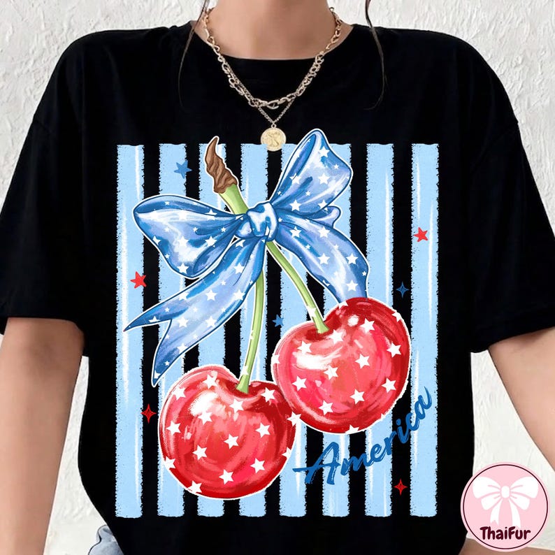 May include: Black t-shirt featuring a patriotic design with two red cherries adorned with white stars. A blue bow with white stars sits above the cherries, set against a blue and black striped background. The word "America" is written in blue.