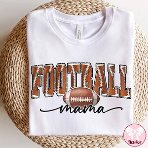 May include: White t-shirt with a football-themed design. The design features the word "FOOTBALL" in brown with white stitching, a brown football, and the word "mama" in a cursive font. The shirt is displayed on a woven surface.