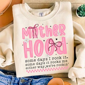 May include: A cream-colored sweatshirt with a pink graphic that says "Mother Hood" with a pink bow and flowers. The text below the graphic says "some days I rock it, some days it rocks me, either way we're rockin'"