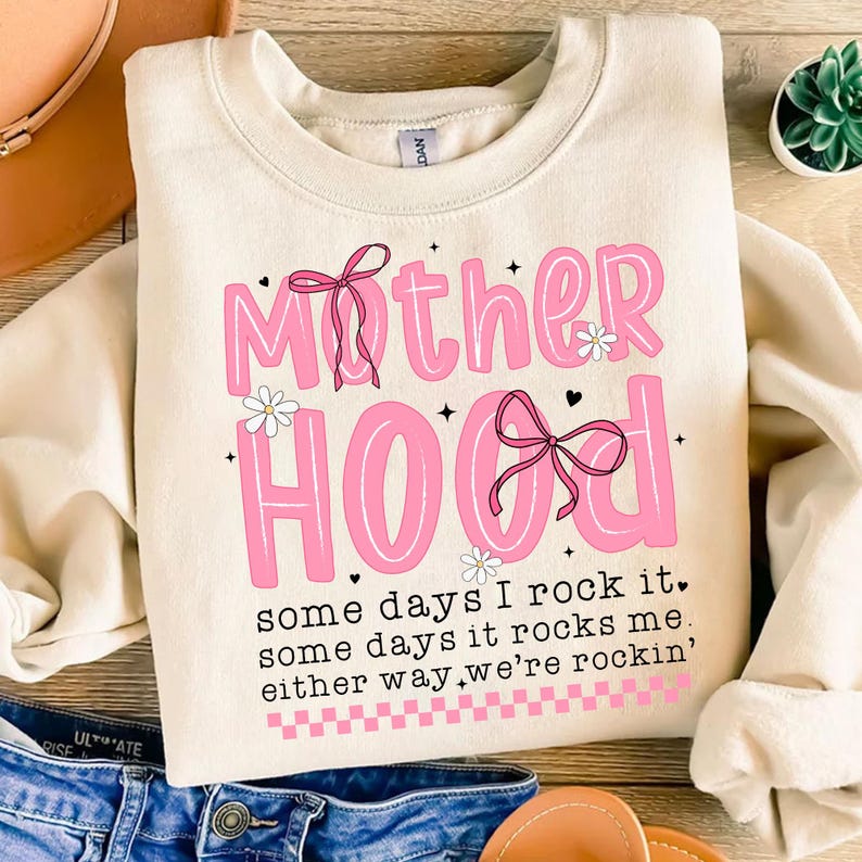 May include: A cream-colored sweatshirt with a pink graphic that says "Mother Hood" with a pink bow and flowers. The text below the graphic says "some days I rock it, some days it rocks me, either way we're rockin'"