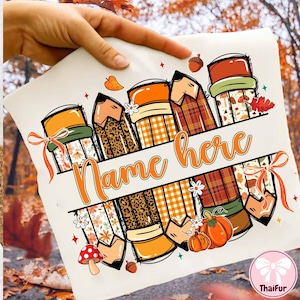May include: A cream-colored pillow with a fall-themed pencil design. The pencils have various patterns, including leopard print, plaid, and floral designs. The text "Name here" is written in orange. The design includes pumpkins, acorns, and leaves.