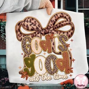 Puede incluir: Tela blanca con la frase "God is Good all the time" en una fuente decorativa. Las letras tienen un estampado de leopardo, acentos marrones y verdes. Un gran lazo con un diseño de estampado de leopardo en la parte superior.