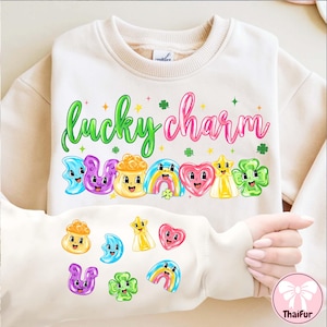 Puede incluir: Sudadera color crema con las palabras "lucky charm" en letras verdes y rosas. Debajo del texto hay amuletos de dibujos animados coloridos, incluyendo una luna sonriente, un pote de oro, un arcoíris, un corazón, una estrella y un trébol. Hay amuletos adicionales en la manga.