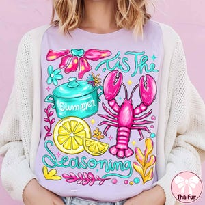 May include: A light purple t-shirt with a colorful graphic design. The design includes a pink lobster, a turquoise pot with the word "Summer", a yellow lemon, and the text "'Tis The Seasoning".