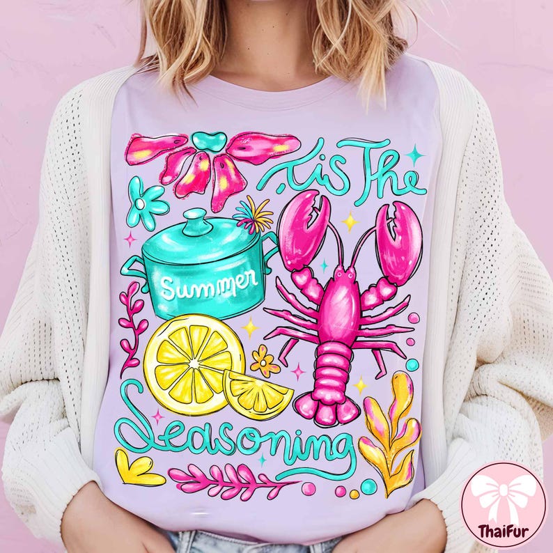 May include: A light purple t-shirt with a colorful graphic design. The design includes a pink lobster, a turquoise pot with the word "Summer", a yellow lemon, and the text "'Tis The Seasoning".