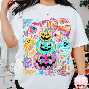 May include: White t-shirt featuring a colorful Halloween-themed graphic. The design includes stacked jack-o'-lanterns, ghosts, bats, and a witch's hat, all in vibrant rainbow hues. A large bow and stars complete the festive look.