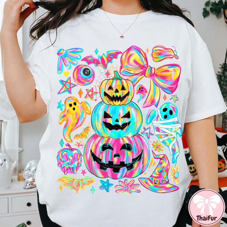 May include: White t-shirt featuring a colorful Halloween-themed graphic. The design includes stacked jack-o'-lanterns, ghosts, bats, and a witch's hat, all in vibrant rainbow hues. A large bow and stars complete the festive look.