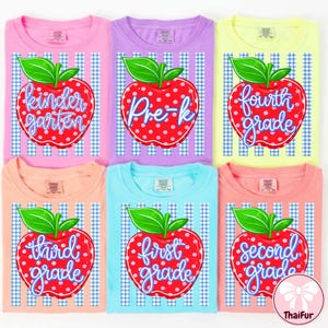 May include: Six colorful t-shirts with school grade level designs. Each shirt features a red apple graphic with a grade level label, such as "Kindergarten" or "First Grade". The shirts are in various colors, including pink, purple, and blue.