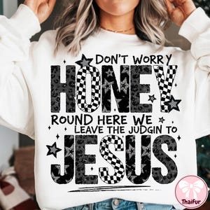 May include: White sweatshirt with the text "DON'T WORRY HONEY ROUND HERE WE LEAVE THE JUDGIN TO JESUS" in black and gray. The word "HONEY" has a checkered pattern and leopard print. The word "JESUS" has a checkered pattern.