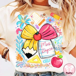 Puede incluir: Camiseta beige con un diseño gráfico colorido. El diseño incluye un lápiz amarillo, un lazo rosa y una manzana roja. El texto "Mrs. Kayce" está escrito en un bloc de notas. Elementos florales y escolares rodean el diseño central.