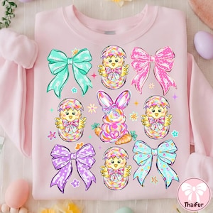 May include: Pink sweatshirt with a colorful Easter-themed design. The design includes bows, chicks hatching from eggs, and a bunny with a carrot. The graphic is in shades of pink, blue, yellow, and purple.