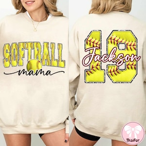 Op de afbeelding: Cr&egrave;mekleurige sweatshirt met de tekst "SOFTBALL mama" en een softbalafbeelding op de voorkant. De achterkant heeft het nummer 19 met een softbalontwerp en de naam "Jackson". Een casual, sportief kledingstuk.