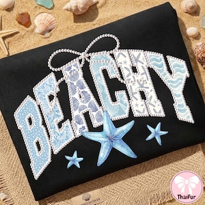 Puede incluir: Toalla de playa negra con la palabra "BEACHY" en blanco, azul y detalles de perlas. Las letras están decoradas con patrones de conchas marinas y olas. Tres estrellas de mar azules están en la toalla.