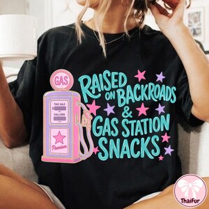 K&ouml;nnte beinhalten: Ein schwarzes Kurzarm-T-Shirt mit einer Grafik einer rosa und lila Vintage-Zapfs&auml;ule. Der Text "RAISED ON BACKROADS & GAS STATION SNACKS" ist in hellblauen Buchstaben gehalten und von kleinen rosa und lila Sternen um das Design herum akzentuiert.