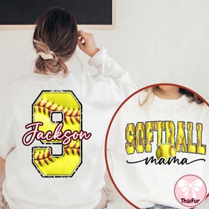 Op de afbeelding: Witte sweatshirt met softbalontwerpen. E&eacute;n ontwerp toont een gele softbal met het nummer 9 en de naam "Jackson" in roze script. Een ander ontwerp zegt "Softball mama" met een softbalafbeelding.