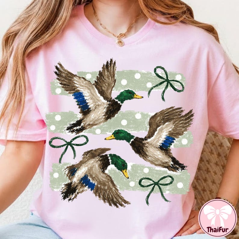 Pu&ograve; includere: Una t-shirt rosa chiaro con una stampa grafica di tre germani reali in volo. Il design include strisce orizzontali a pennellate verde salvia, pois bianchi e fiocchi disegnati a linee verde scuro sparsi sulla parte anteriore.