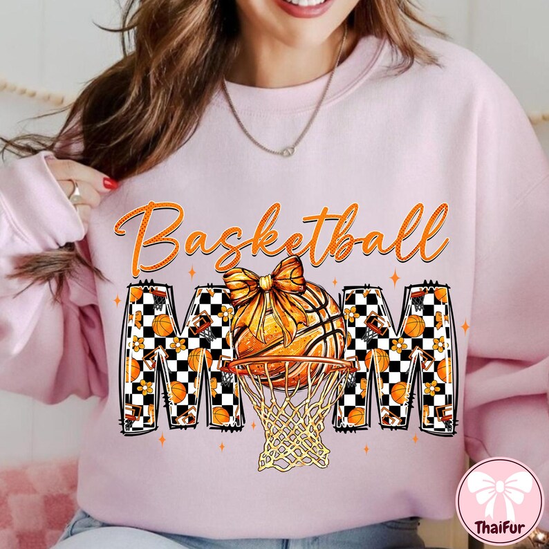 May include: Light pink sweatshirt with the words "Basketball Mom" in orange script. The word "MOM" is designed with a basketball, net, and checkered pattern. A basketball with a bow is in the center.